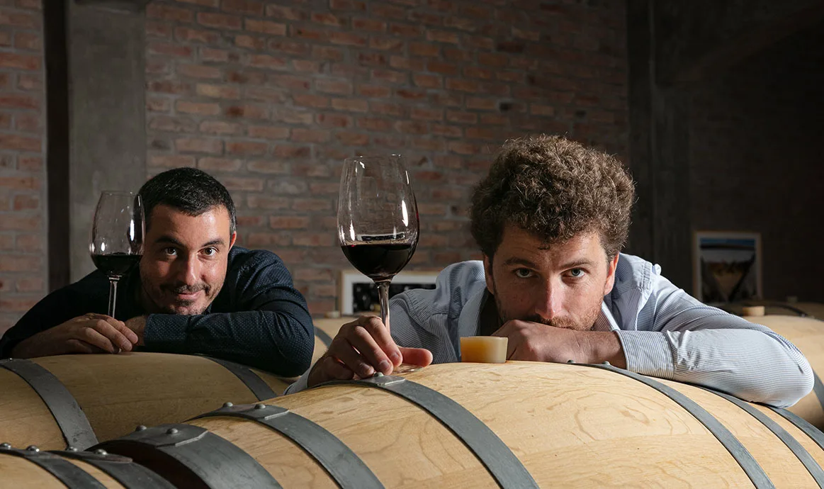 Quentin y Thibault con copas de vino sobre las barricas